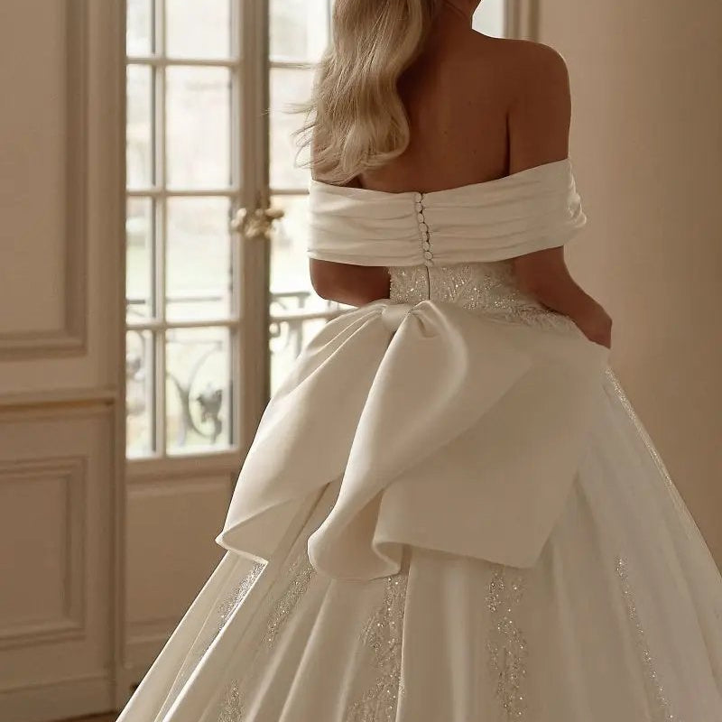 Off-shoulder satin bridal gown with draped bow and beaded waist detailing